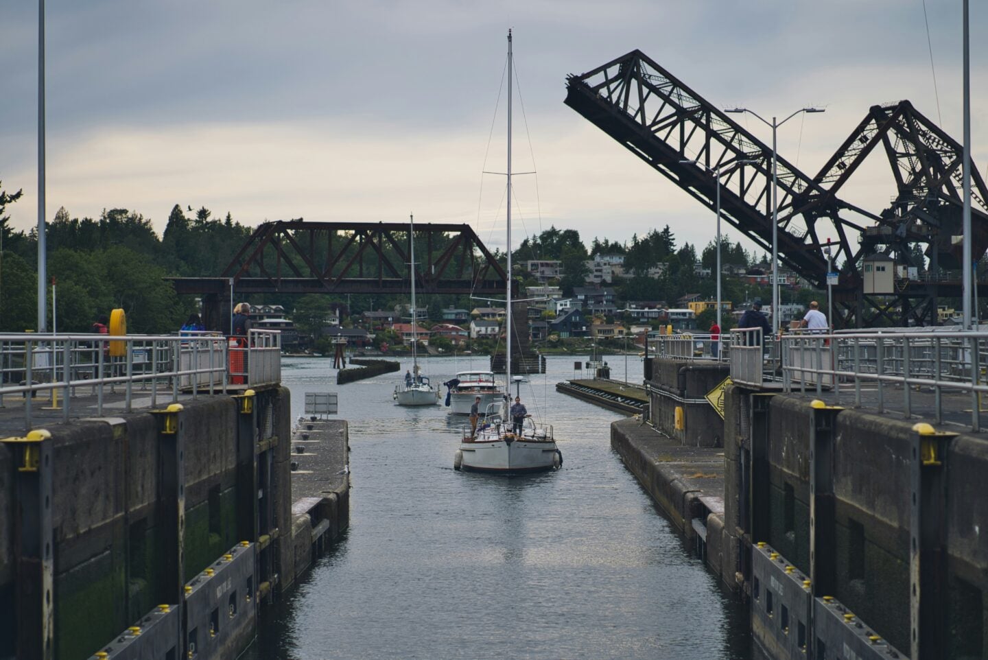 Sailboats pass through the Ballard Locks in Seattle as raised drawbridges and a railroad bridge frame the waterway.