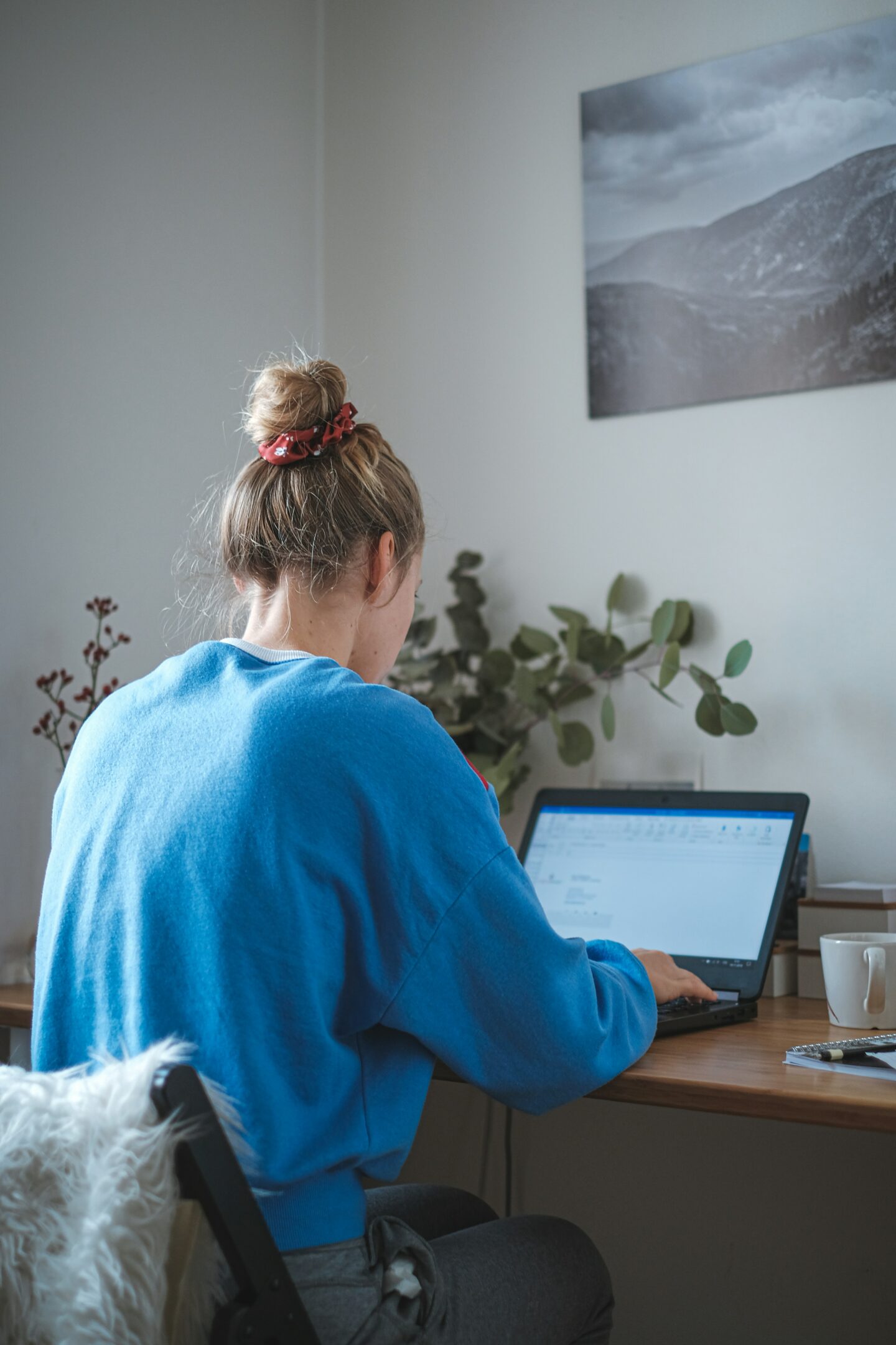 A person in a blue sweater working on a laptop at a wooden desk in a home office, with plants, a notebook, and a mug nearby, showing a remote work setup.