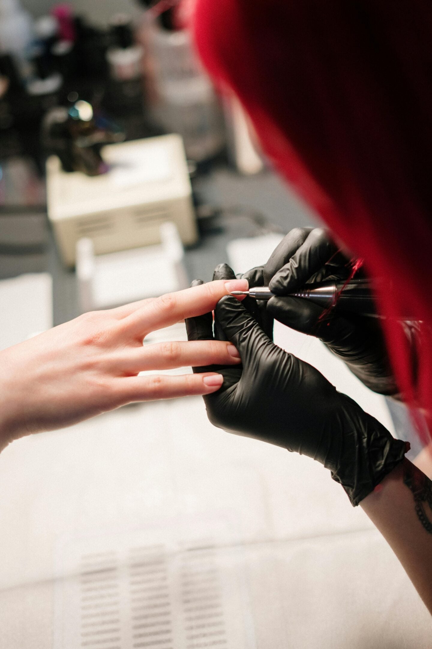 A nail technician wearing black gloves shaping a client’s nail with electric nail drill during a manicure.