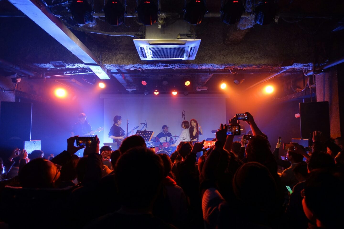 A crowded indoor concert where audience members hold up phones to record the band performing on stage under colorful lights.