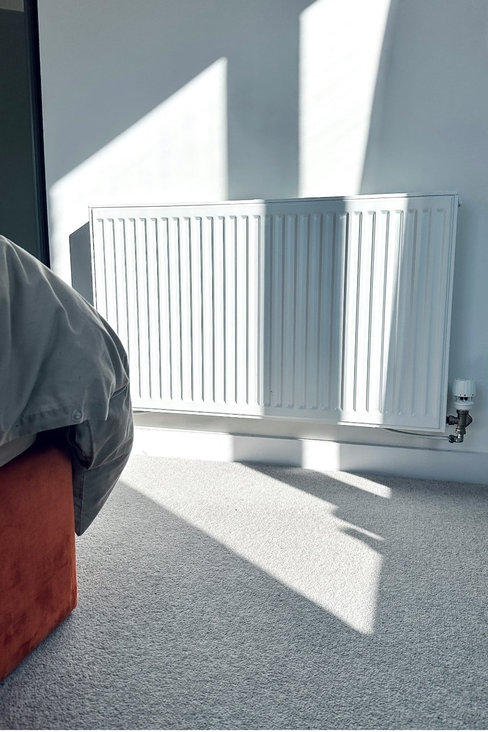 Sunlight streaming across a carpeted bedroom floor toward a white radiator mounted on the wall, highlighting a calm, minimalist interior.