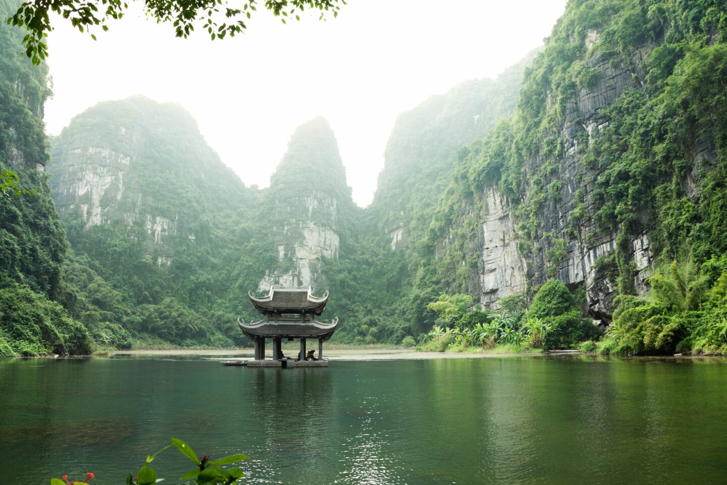 Pagoda pavilion reflected on calm water surrounded by limestone karst mountains in Tràng An, Ninh Bình, Vietnam.