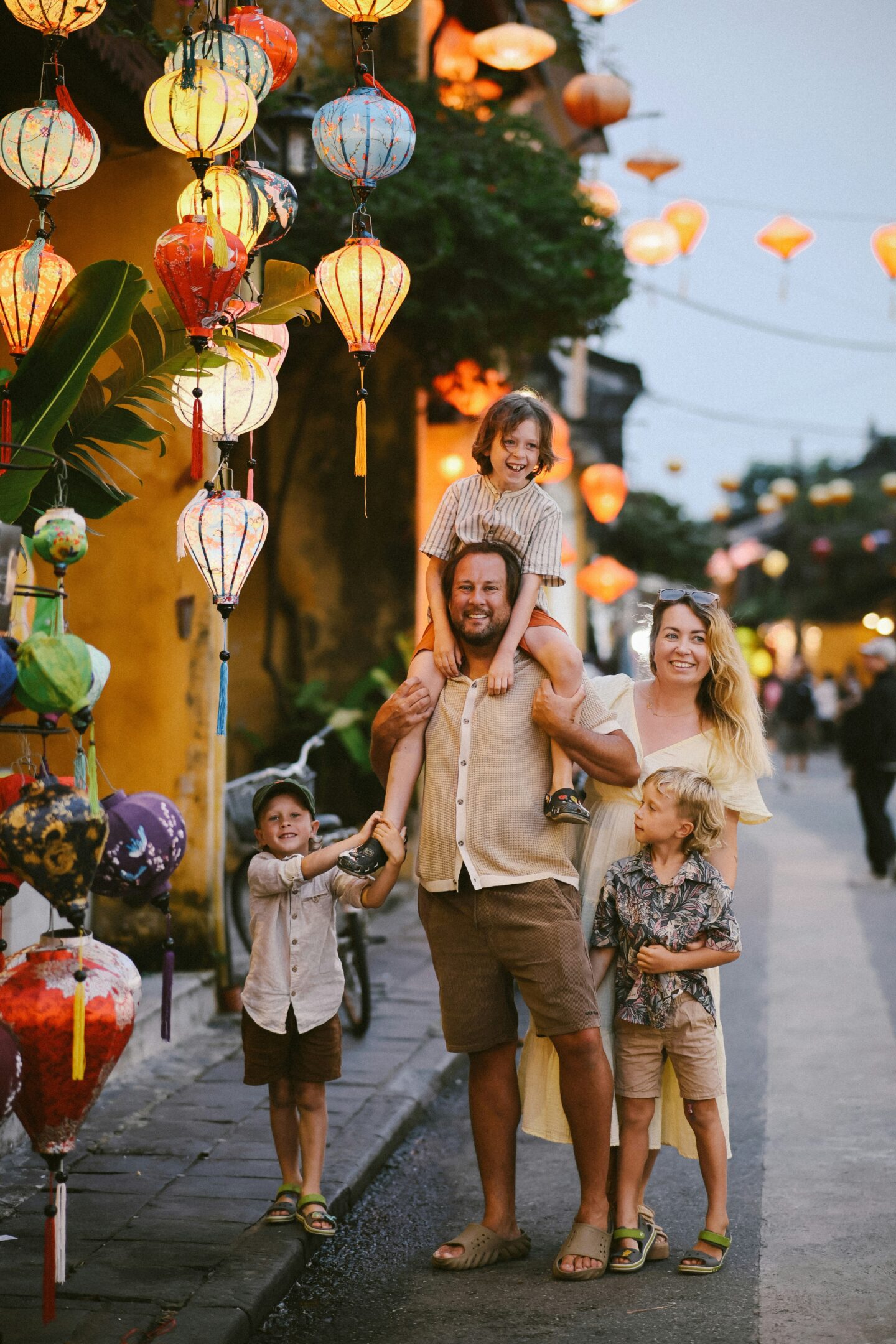 A family of five walking together down a lantern-lit street at dusk, with a child sitting on a parent’s shoulders, capturing a warm family travel moment in a lively destination.
