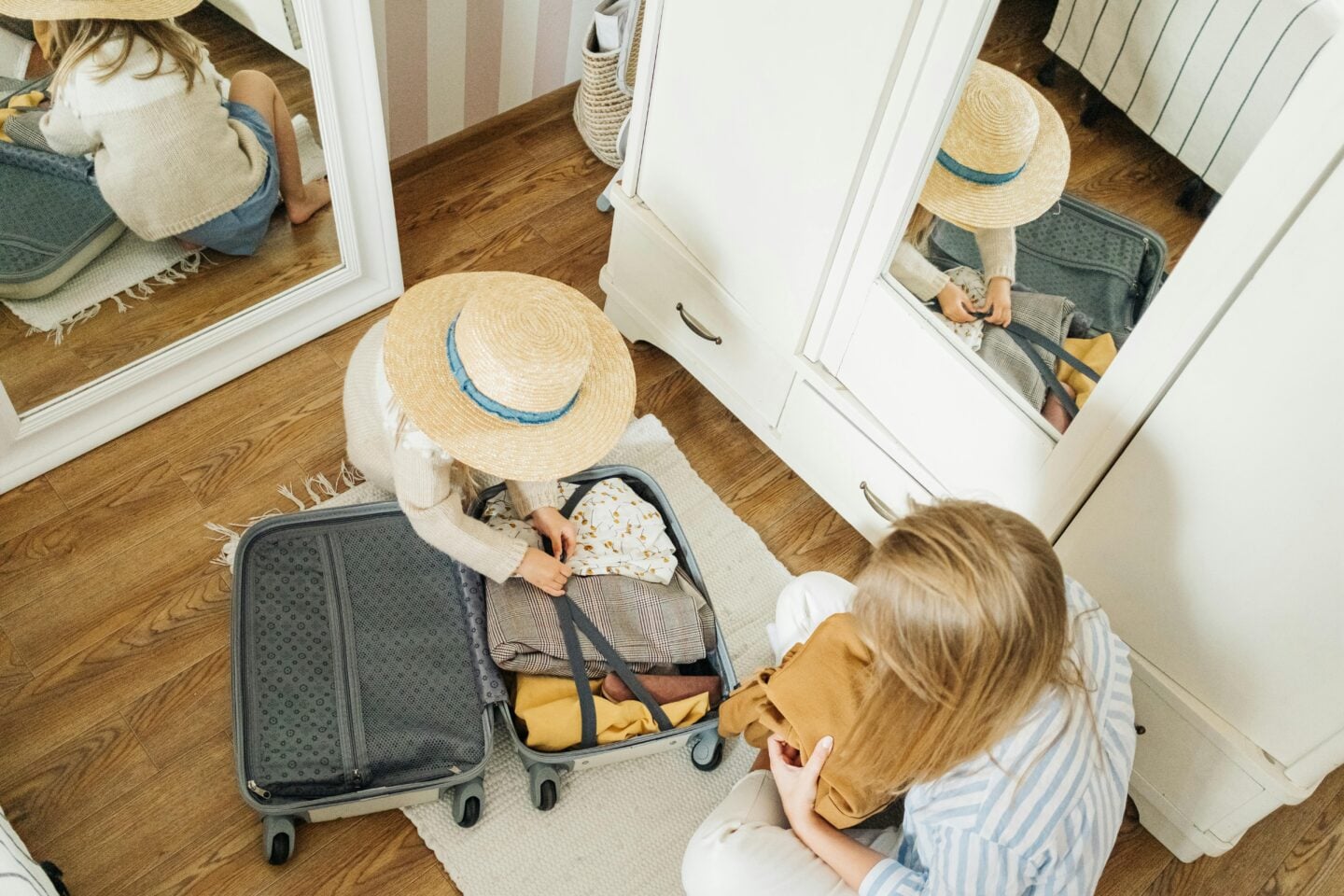 A parent and child packing clothes together into an open suitcase on a bedroom floor, with a mirror reflecting the scene, suggesting family travel preparation at home.