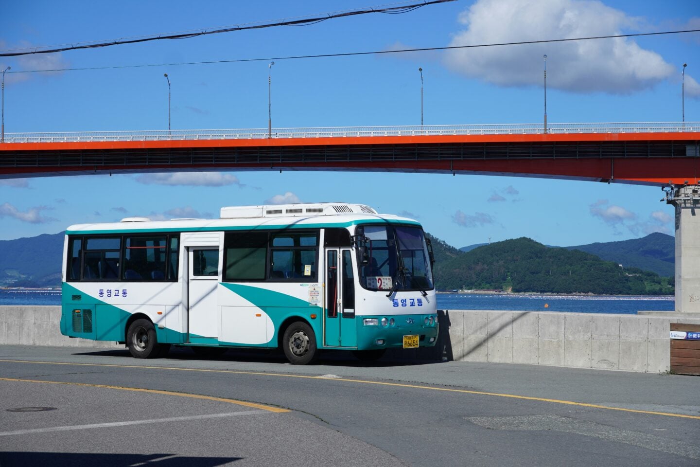 A small city bus parked beside a road near the water, with a bridge overhead and hills in the background, representing local public transportation during travel.