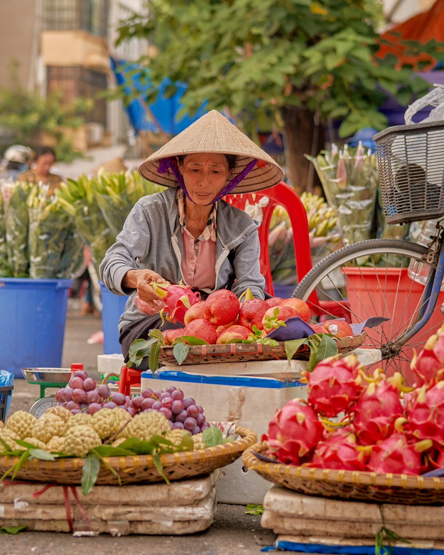 A street vendor wearing a traditional conical hat arranging fresh fruit at an outdoor market stall, with baskets of dragon fruit and other produce displayed in front.