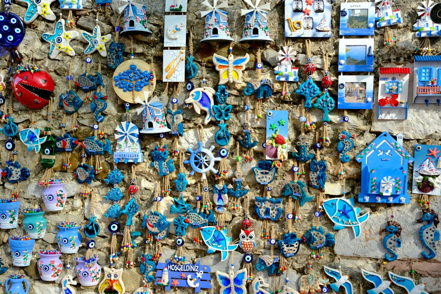 A stone wall covered with colorful handmade souvenirs, including evil eye charms, fish, houses, and nautical decorations, commonly sold in tourist areas.