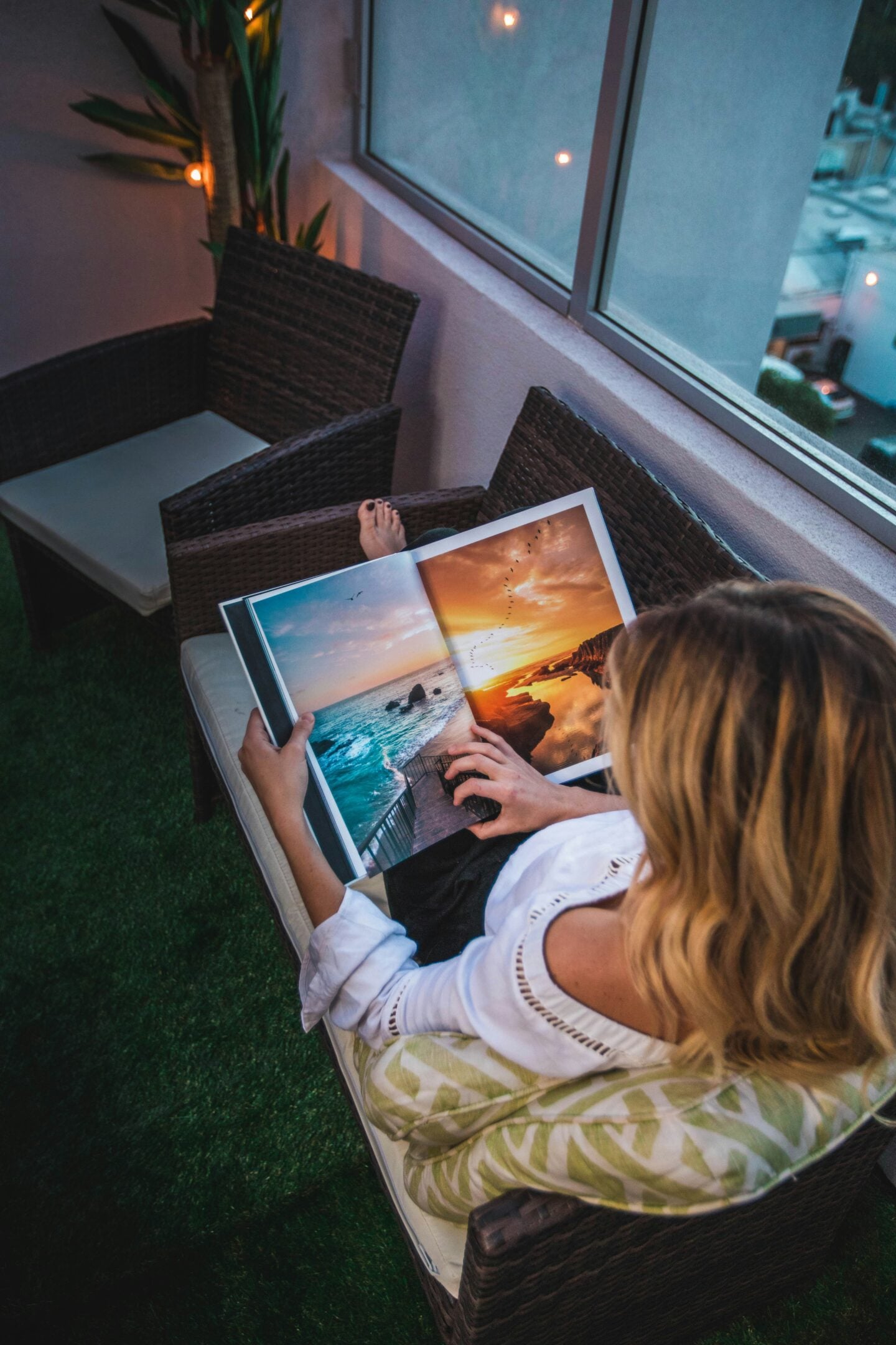 Person sitting on a balcony chair looking through a travel photo book opened to ocean and sunset landscapes, with city lights visible outside.