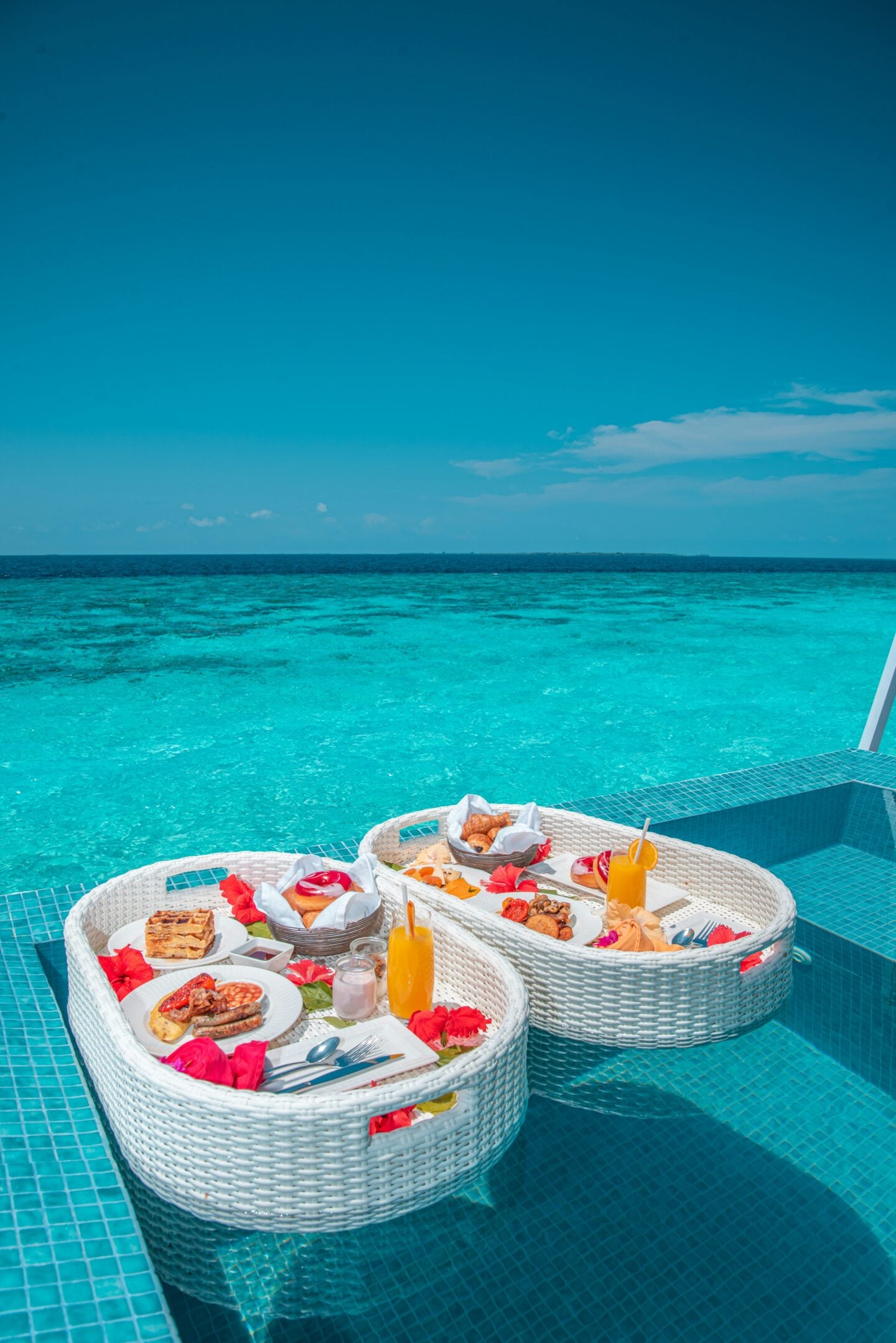 A luxurious floating breakfast tray set in a turquoise pool overlooking the ocean, filled with pastries, fruit, and drinks.
