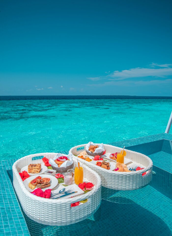 A luxurious floating breakfast tray set in a turquoise pool overlooking the ocean, filled with pastries, fruit, and drinks.