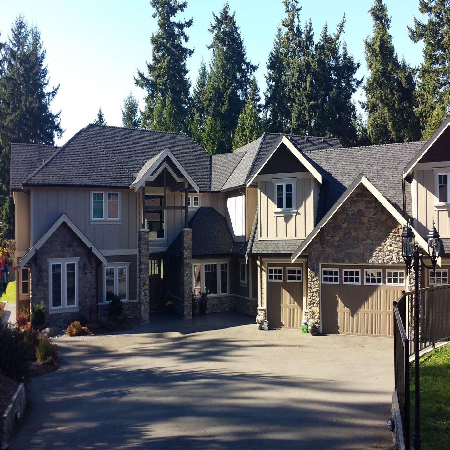 Large suburban house with stone and siding exterior, a wide driveway, and tall evergreen trees in the background on a clear day.