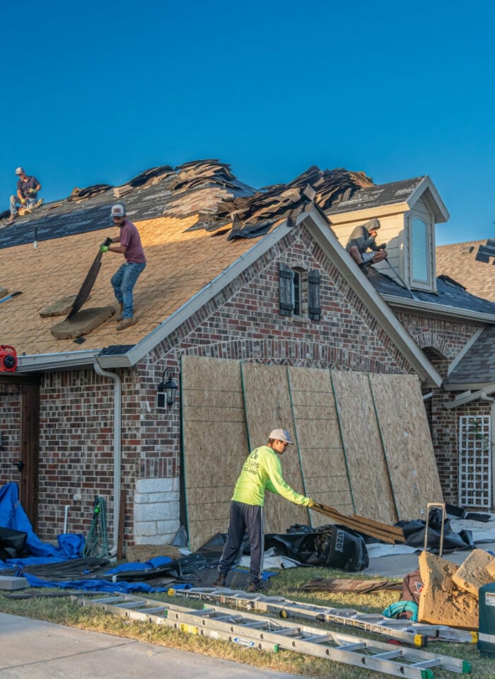 Roofing contractors working on a residential home during a full roof replacement, with old shingles removed and tools and materials spread across the driveway.