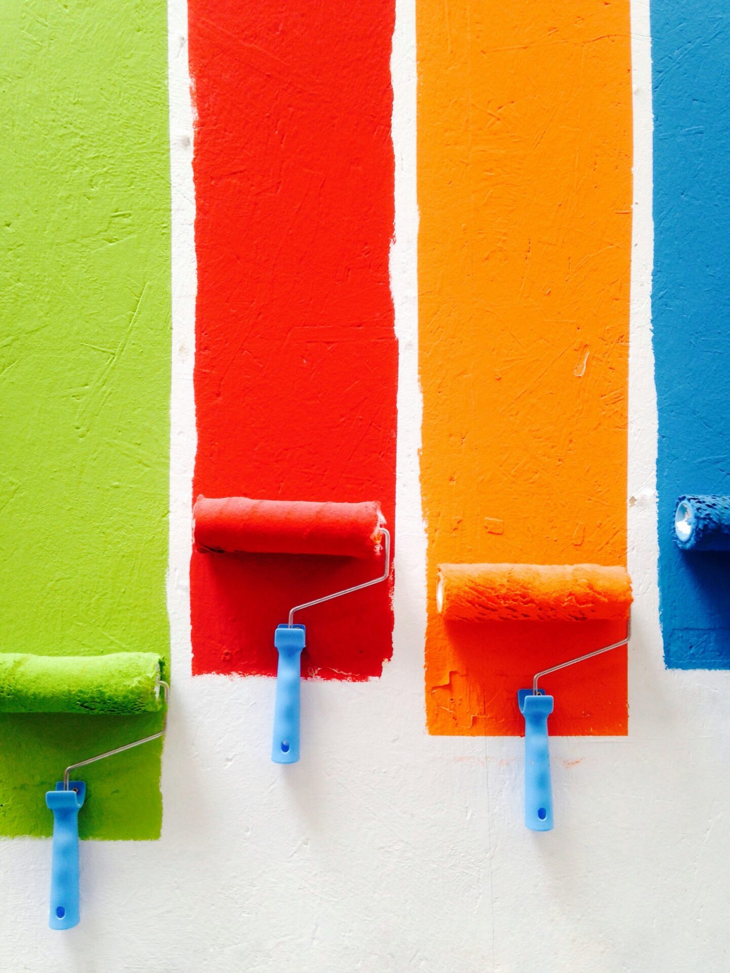 Paint rollers applying bright green, red, orange, and blue paint in vertical stripes on a white wall, showing color options for an exterior painting update.