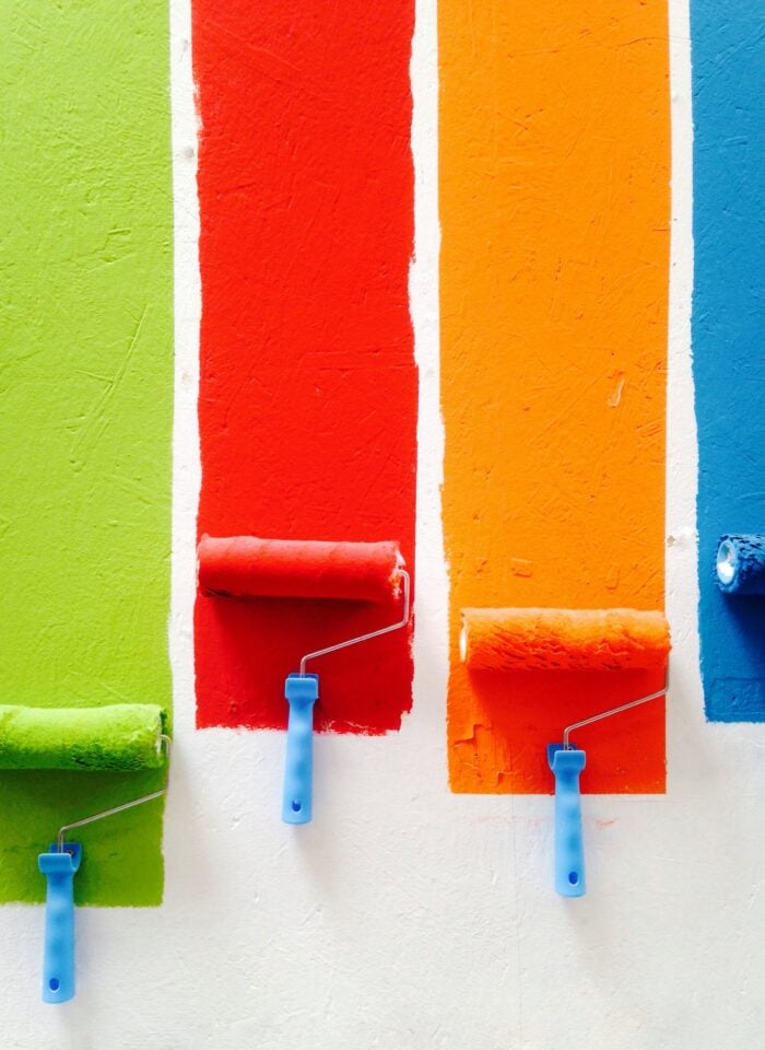 Paint rollers applying bright green, red, orange, and blue paint in vertical stripes on a white wall, showing color options for an exterior painting update.