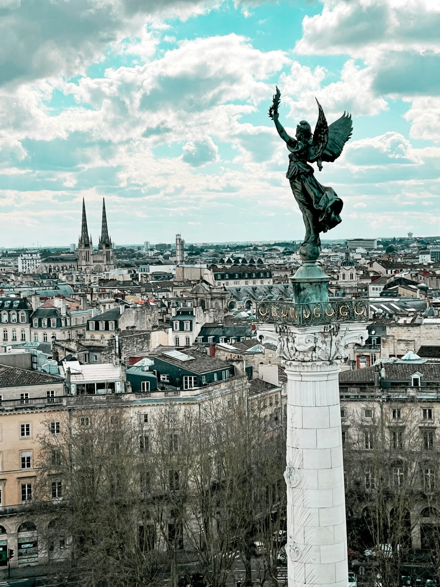 The winged bronze statue crowning the Monument aux Girondins rises above the Bordeaux skyline under a partly cloudy sky.