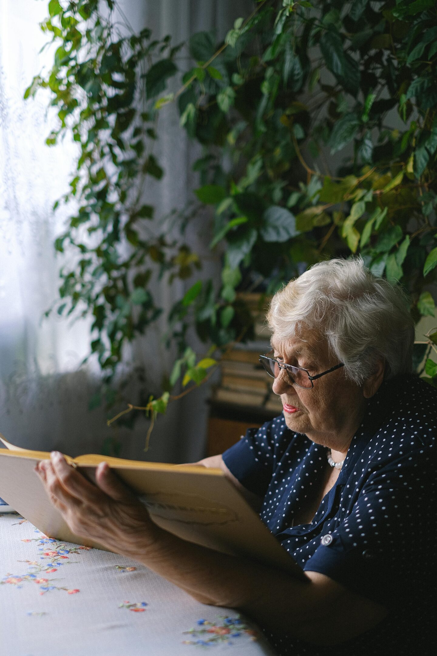 Elderly woman sitting at a table and reading a document in a quiet, plant-filled room at home
