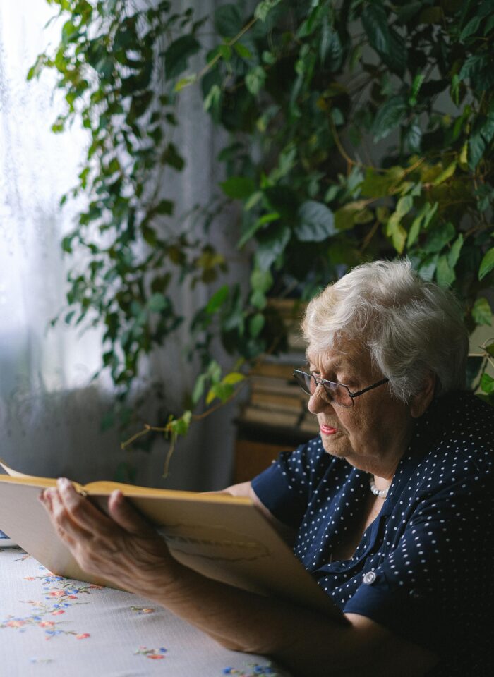 Elderly woman sitting at a table and reading a document in a quiet, plant-filled room at home