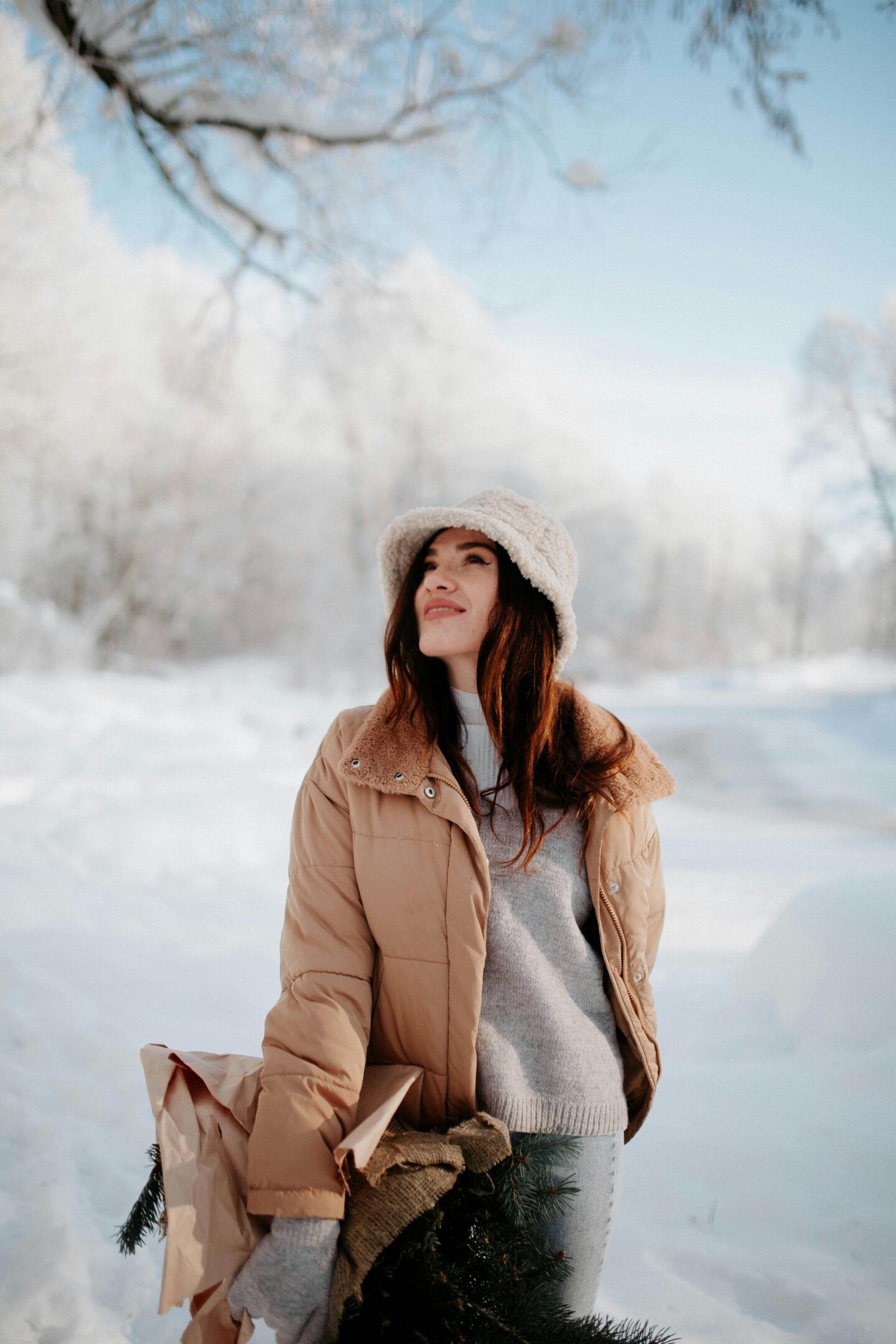 A woman dressed in a beige winter coat and knit hat smiling while standing in a snowy outdoor landscape.