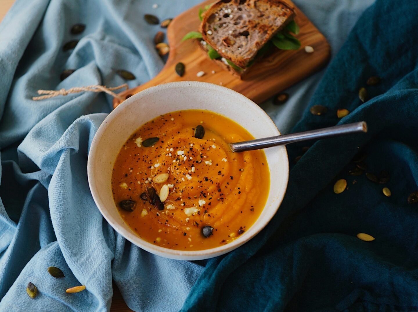 A bowl of creamy pumpkin soup topped with seeds and spices, served alongside a sandwich on a wooden board with blue fabric underneath.