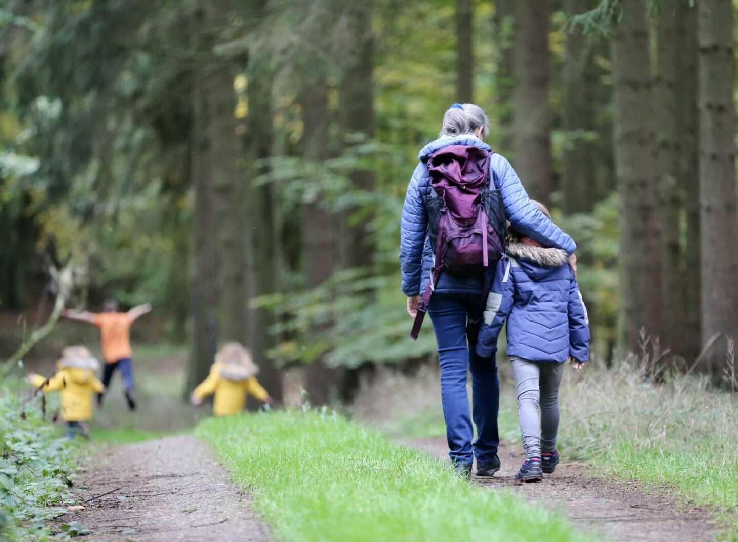 A woman walks through a forest trail with a child at her side, both dressed in jackets and backpacks, while other children run playfully ahead among the trees.