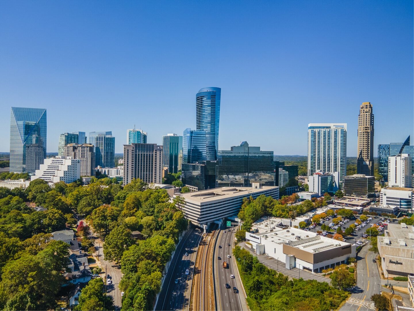 Aerial view of the Atlanta skyline with modern high-rise buildings, tree-lined streets, and blue sky.