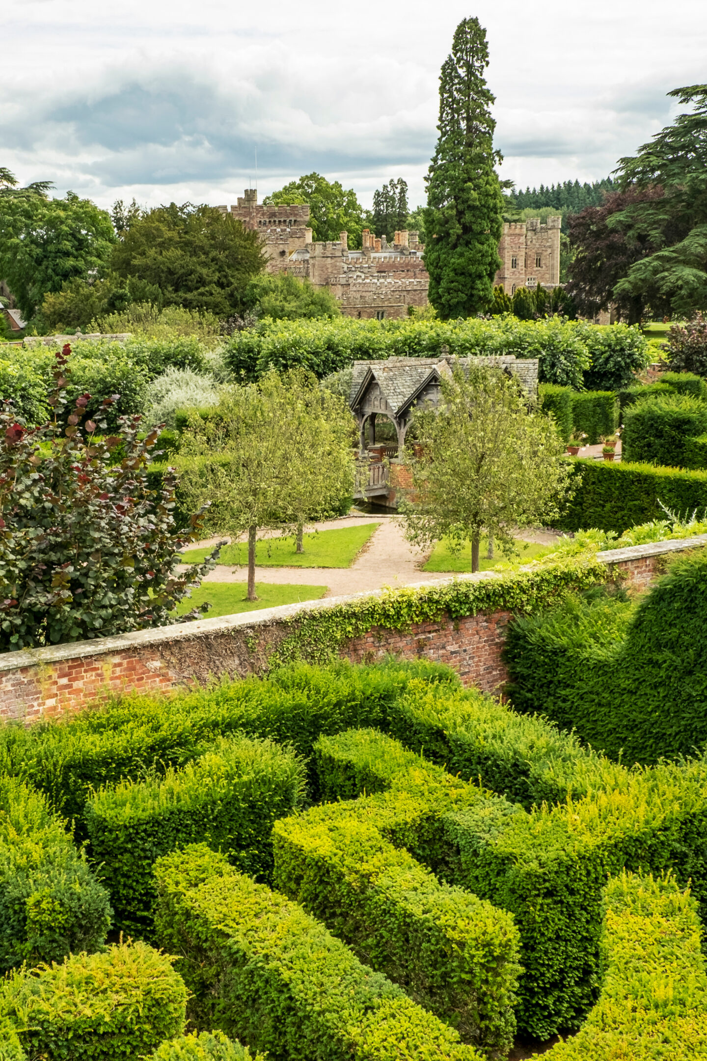 A view over the Hampton Court Palace hedge maze and surrounding formal gardens, with neatly trimmed green hedges, pathways, and a small wooden shelter, and the palace’s brick towers visible among the trees in the background.