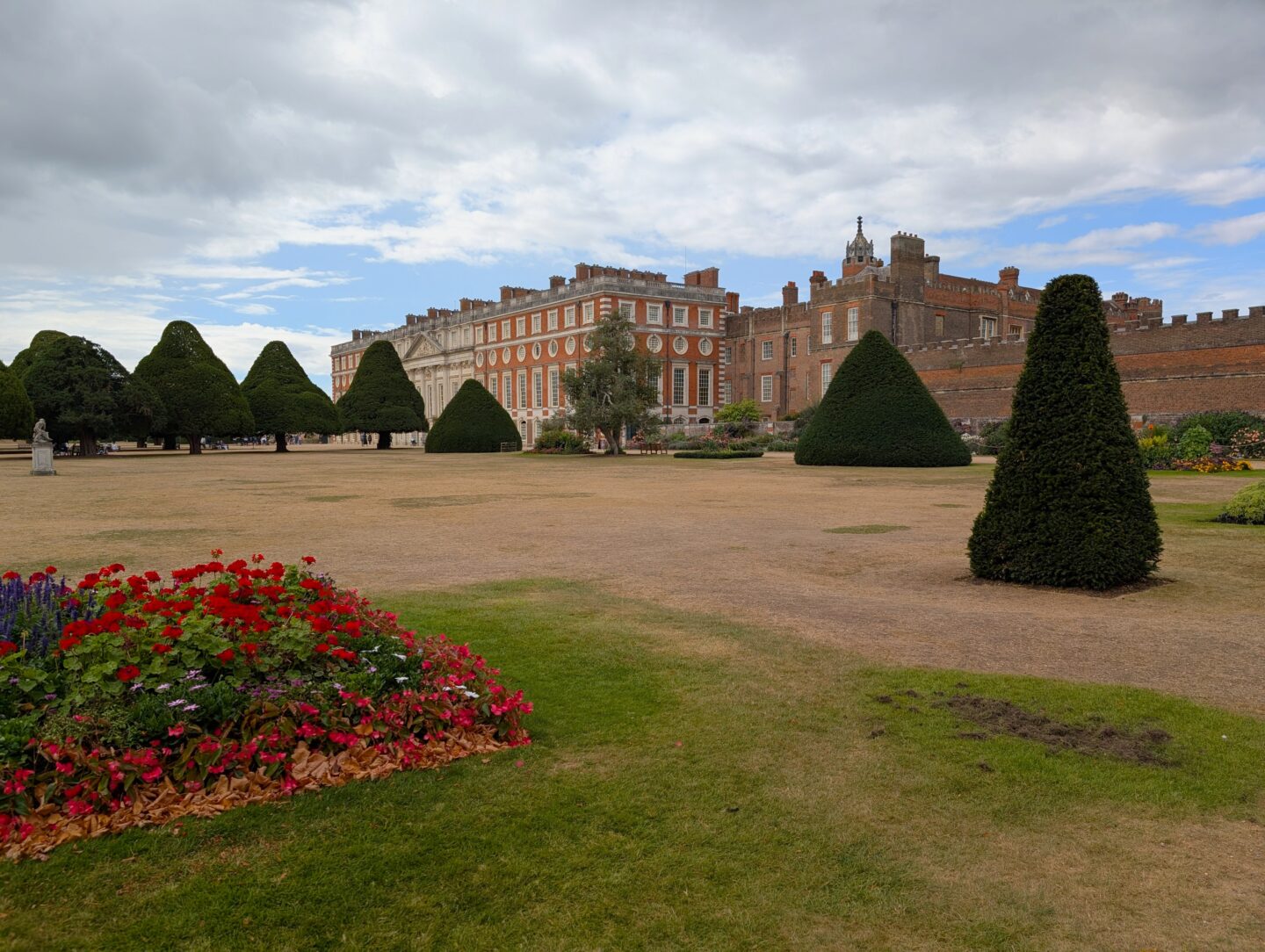 Manicured palace gardens with shaped yew trees and flower beds leading toward the red-brick and stone façade of Hampton Court Palace beneath a cloudy sky.