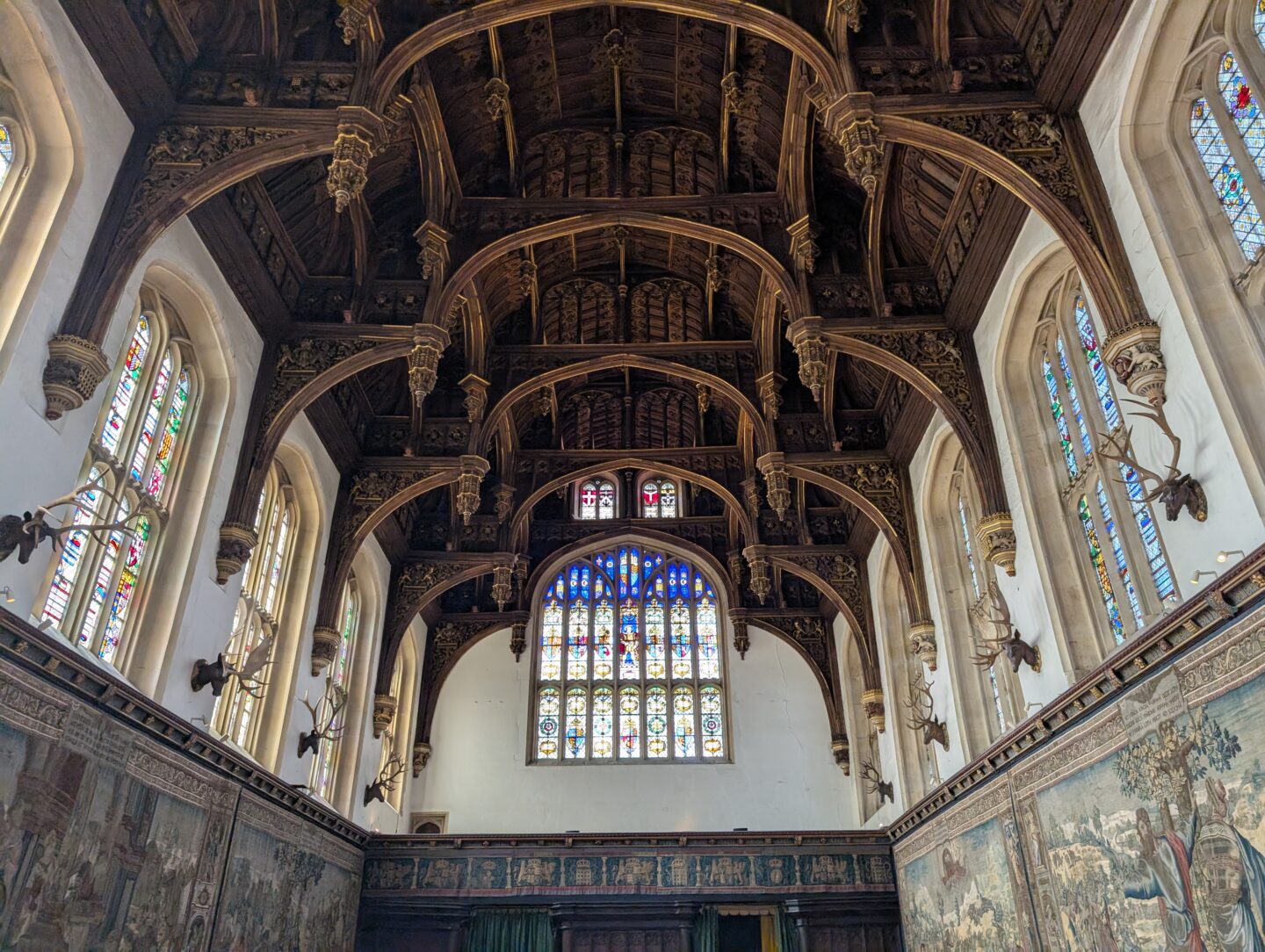The Great Hall at Hampton Court Palace with an intricately carved hammerbeam wooden ceiling, stained glass windows, and tapestry-covered walls hung with antlers.