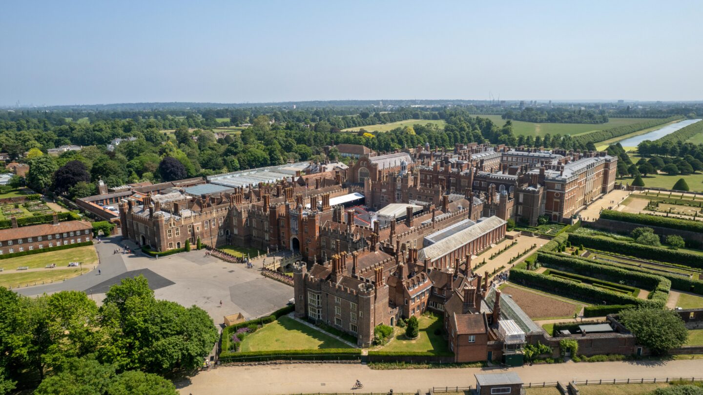 Panoramic aerial view of Hampton Court Palace surrounded by formal gardens, courtyards, and parkland, with the River Thames visible in the background.