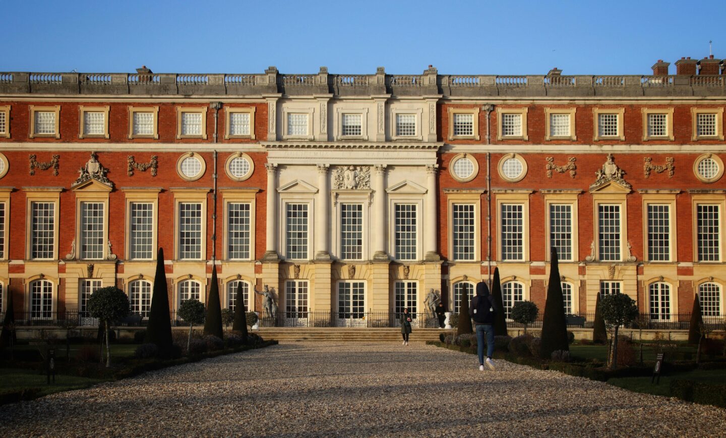 The grand Baroque south front of Hampton Court Palace featuring tall sash windows, ornamental stonework, and a gravel path lined with trimmed topiary trees.