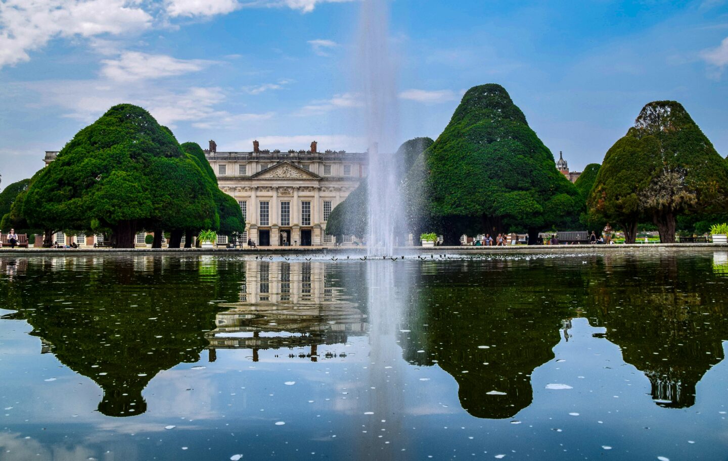 A large fountain spraying water in the center of a reflective pond at Hampton Court Palace Gardens, framed by neatly sculpted yew trees with the grand Baroque palace façade in the background under a blue sky