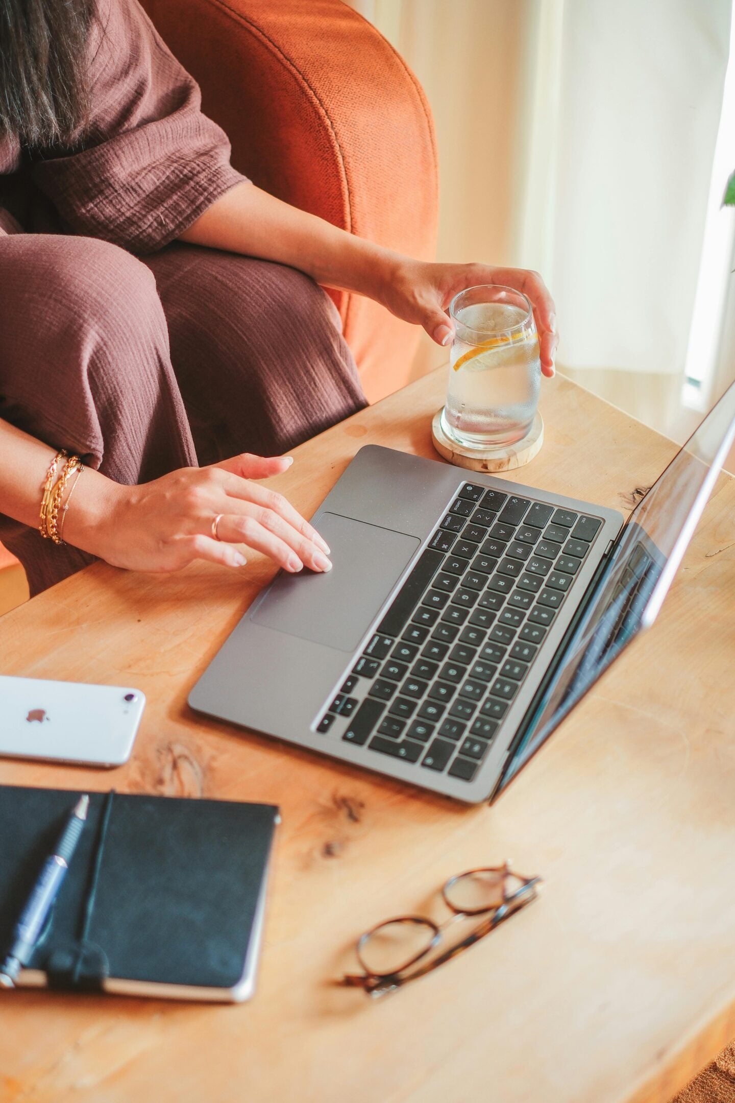 A woman working on a laptop at a wooden table while holding a glass of lemon water, with a phone, notebook, pen, and glasses nearby.
