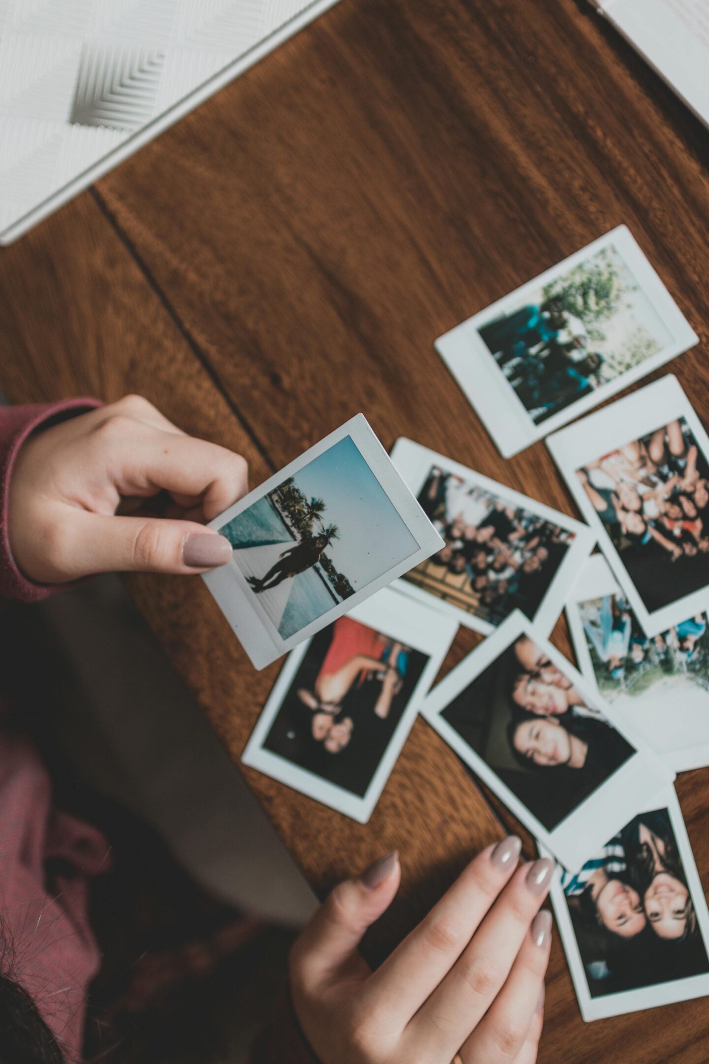 A person sorting through a scattered collection of instant photos on a wooden table, holding one picture featuring a sunny outdoor scene with palm trees.