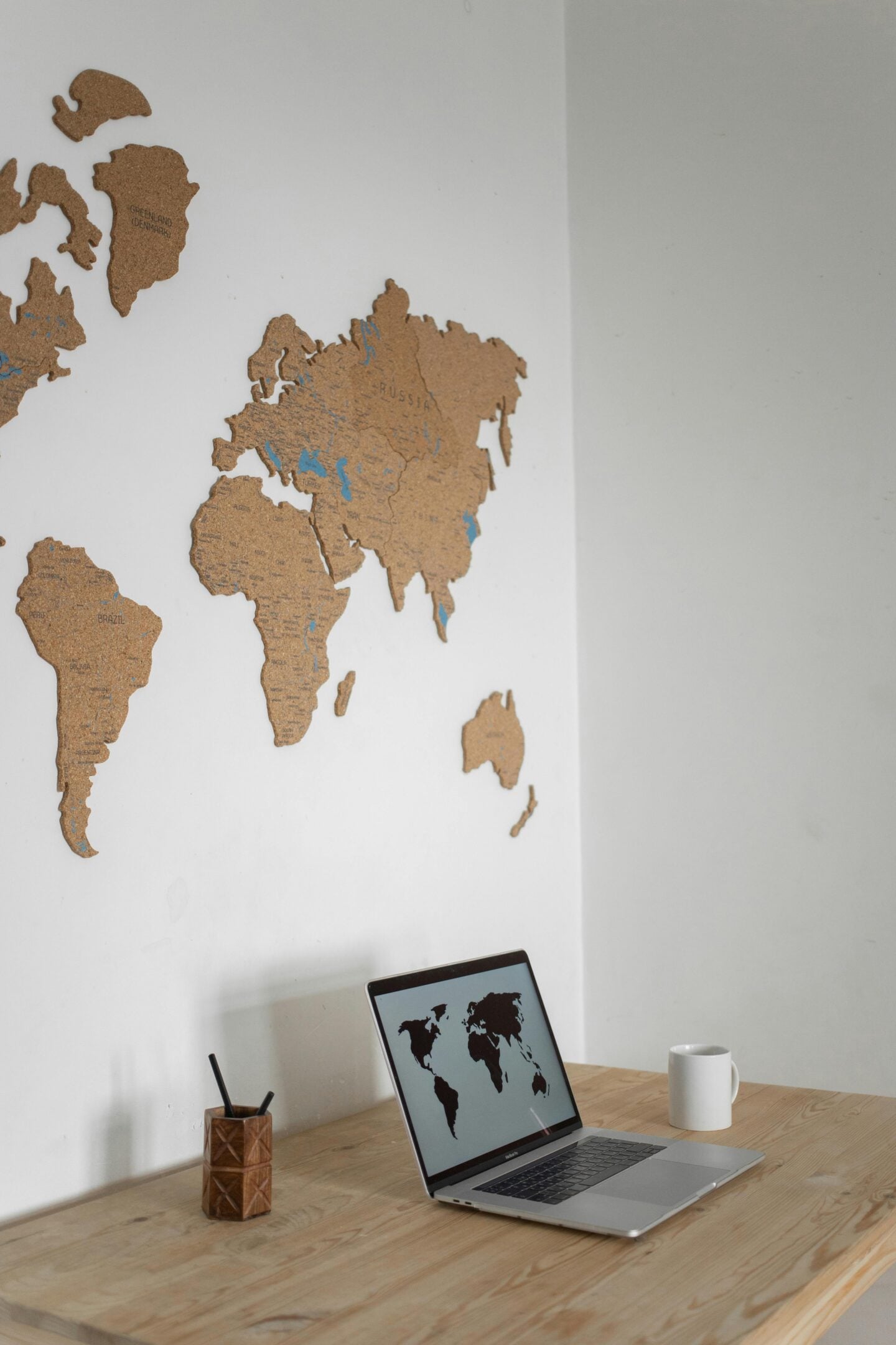 A minimalist workspace with a cork world map on the wall, a laptop on a wooden desk, and a mug and pencil holder beside it.