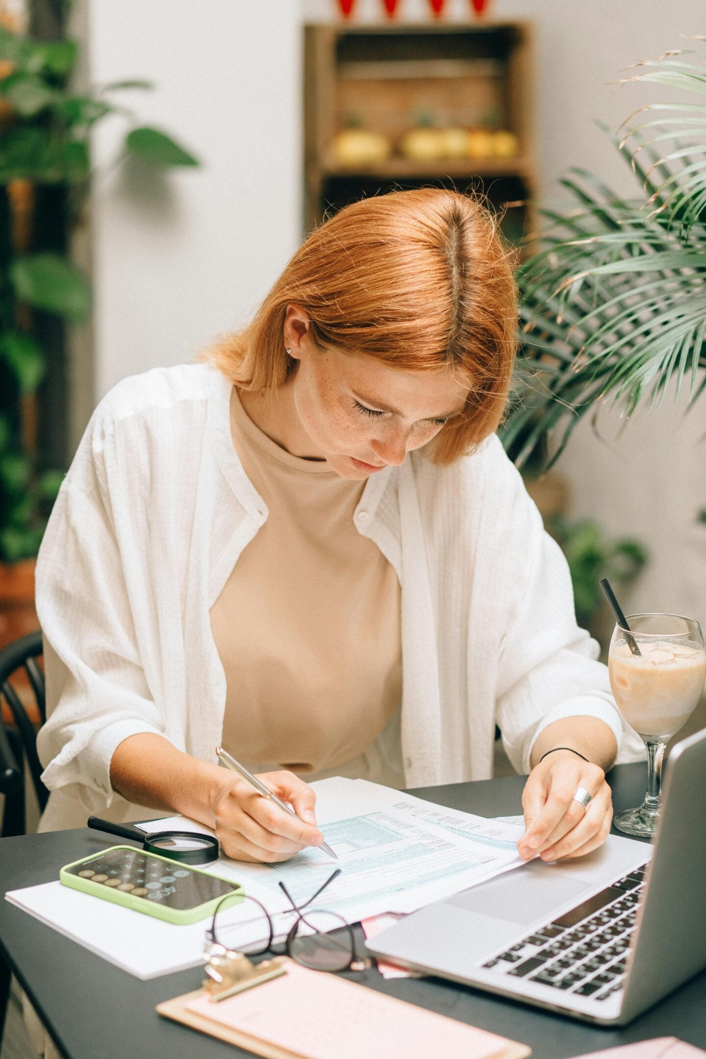 Woman sitting at a desk reviewing financial documents, writing on paperwork next to a laptop, calculator, and iced coffee.
