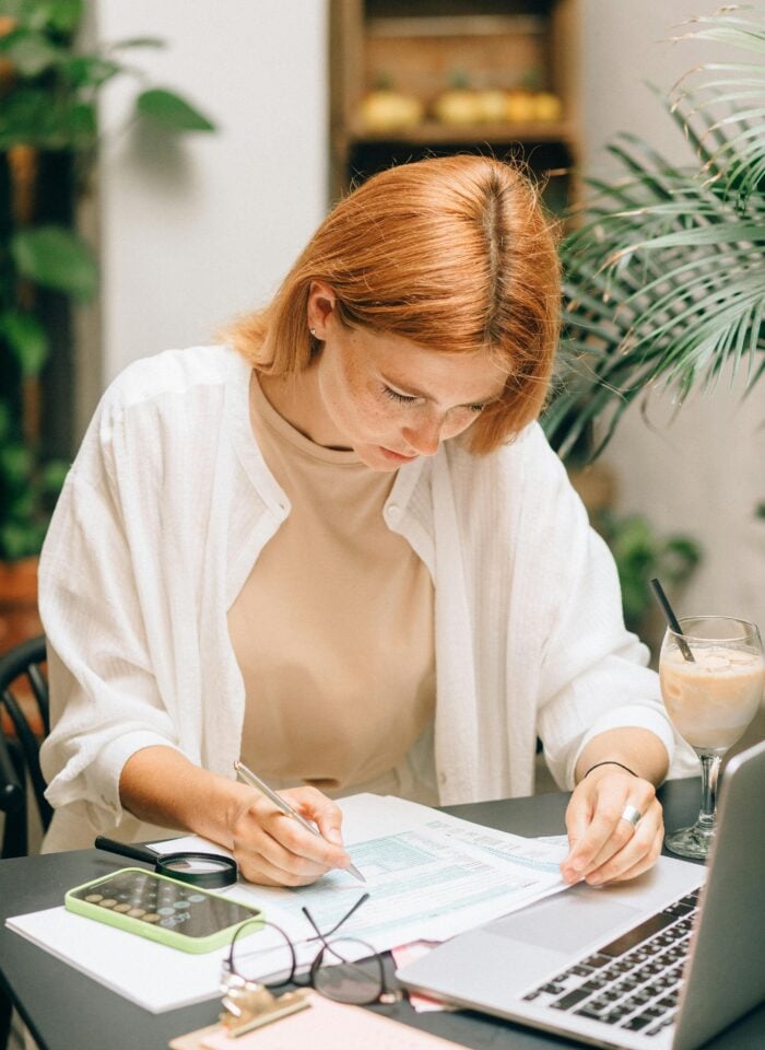 Woman sitting at a desk reviewing financial documents, writing on paperwork next to a laptop, calculator, and iced coffee.