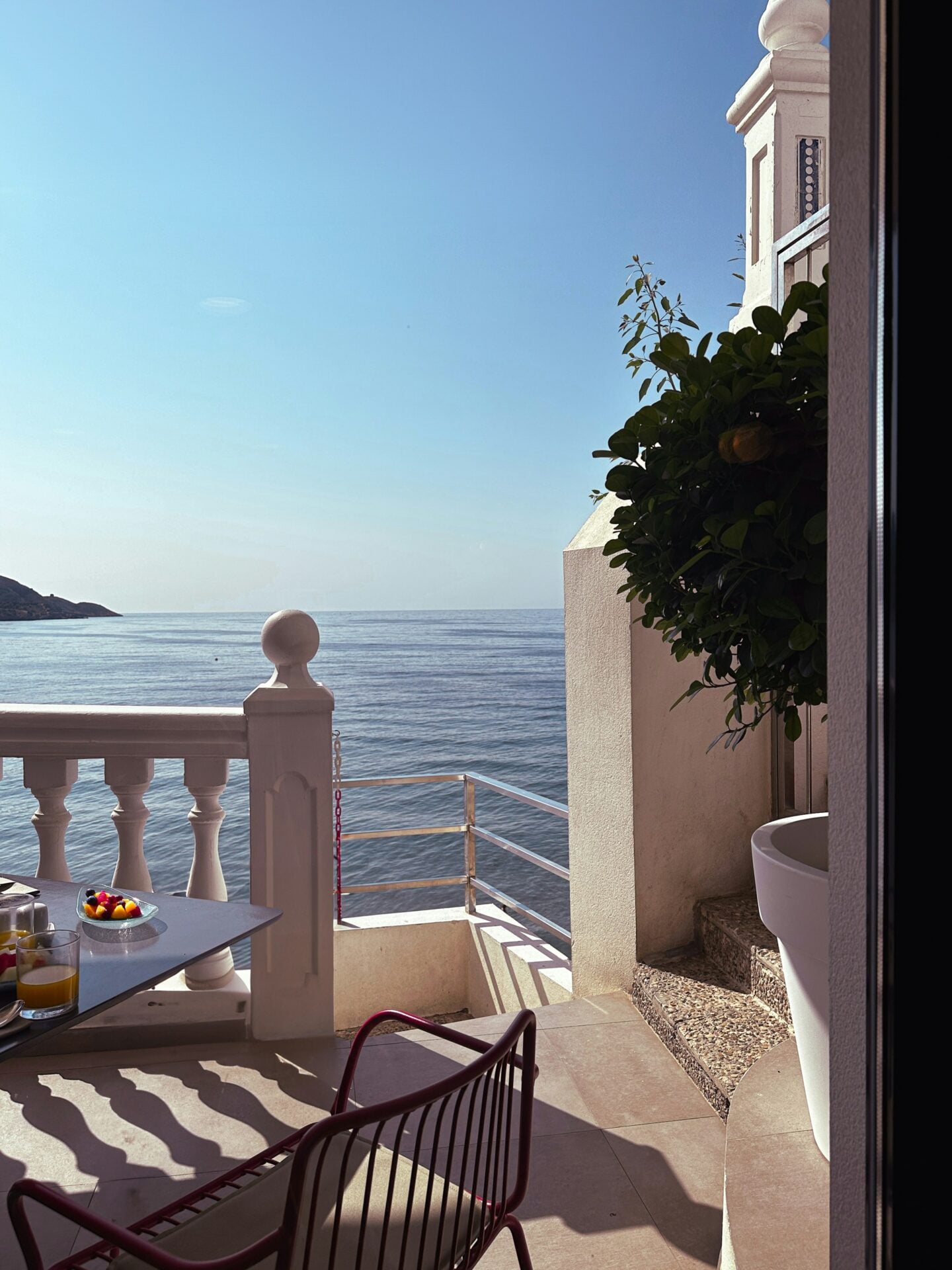 Sunlit seaside terrace in Spain, overlooking calm blue Mediterranean waters, with white balustrades, a small breakfast table, and coastal architecture framing the ocean view.