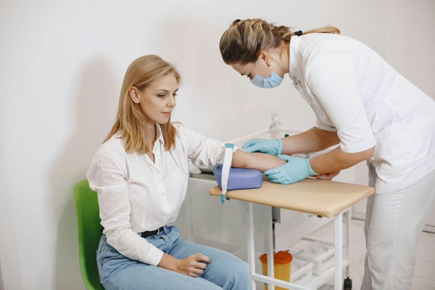A healthcare professional wearing gloves and a face mask prepares to take a blood sample from a seated patient. The patient rests her arm on a cushioned support while the nurse gently holds her forearm in a clean, clinical setting. The scene conveys routine medical care, calm interaction, and professional attention to patient comfort.