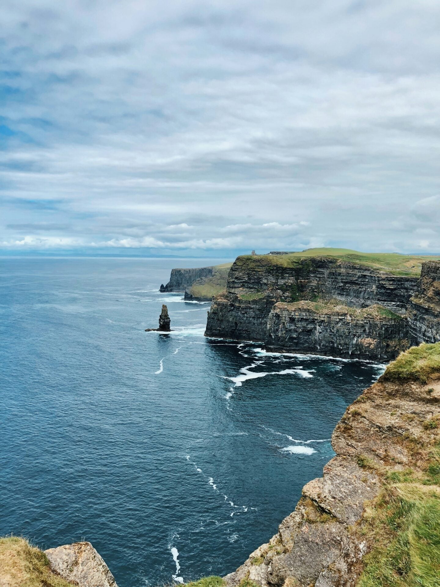 The Cliffs of Moher in Ireland, towering above the Atlantic Ocean with rugged, layered rock faces, grassy cliff tops, waves crashing below, and an overcast sky stretching across the horizon.