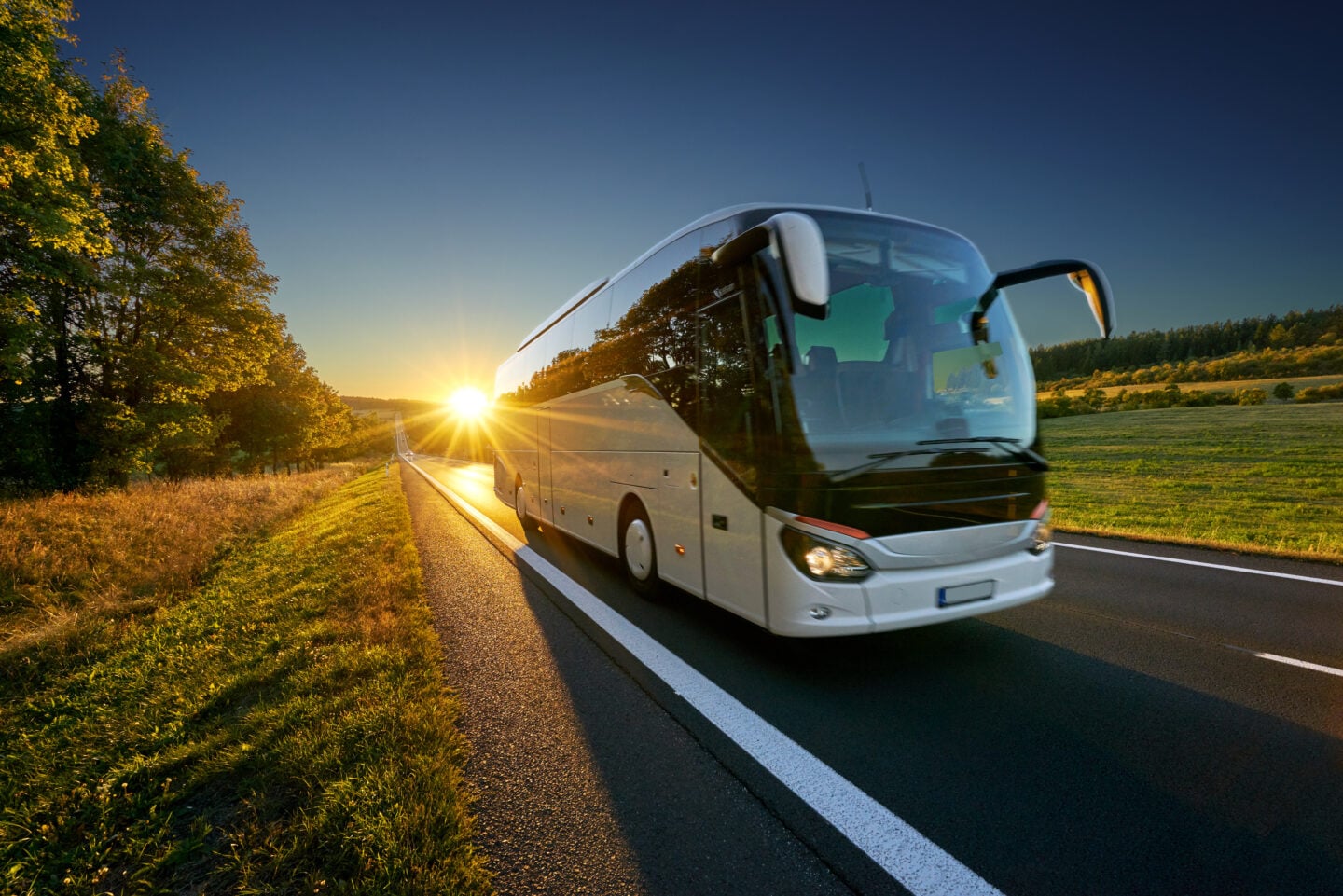 A white coach bus traveling along a rural road at sunset, with golden sunlight, green fields, and trees lining the roadside.