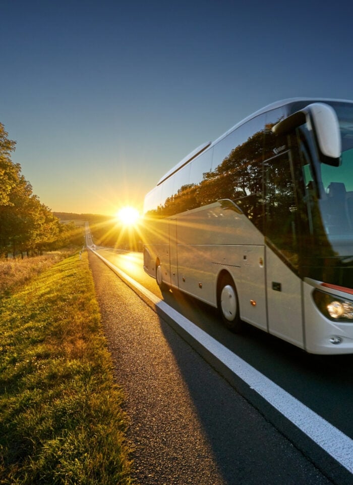 A white coach bus traveling along a rural road at sunset, with golden sunlight, green fields, and trees lining the roadside.