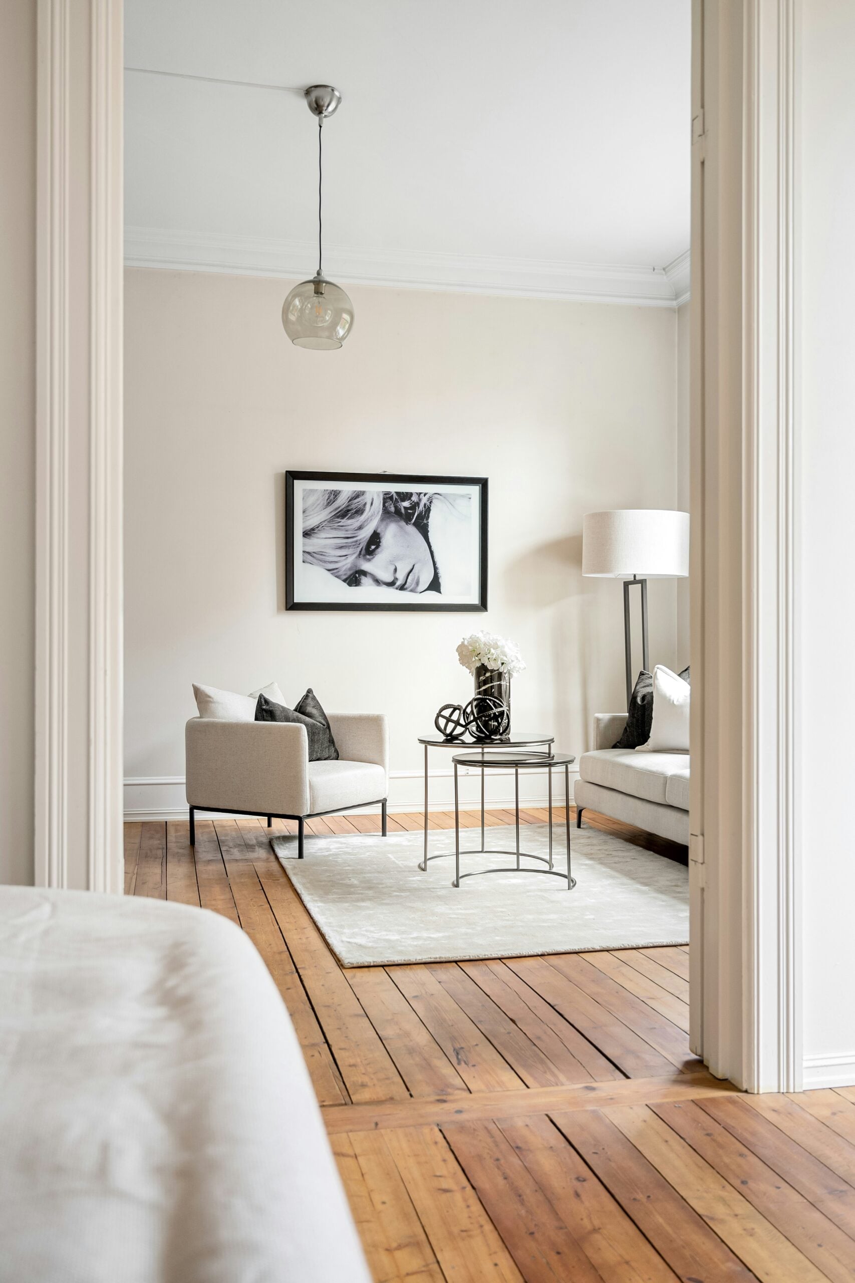Minimalist living room with light neutral armchairs, a small round coffee table, a soft area rug, and warm wooden floors, viewed through a doorway
