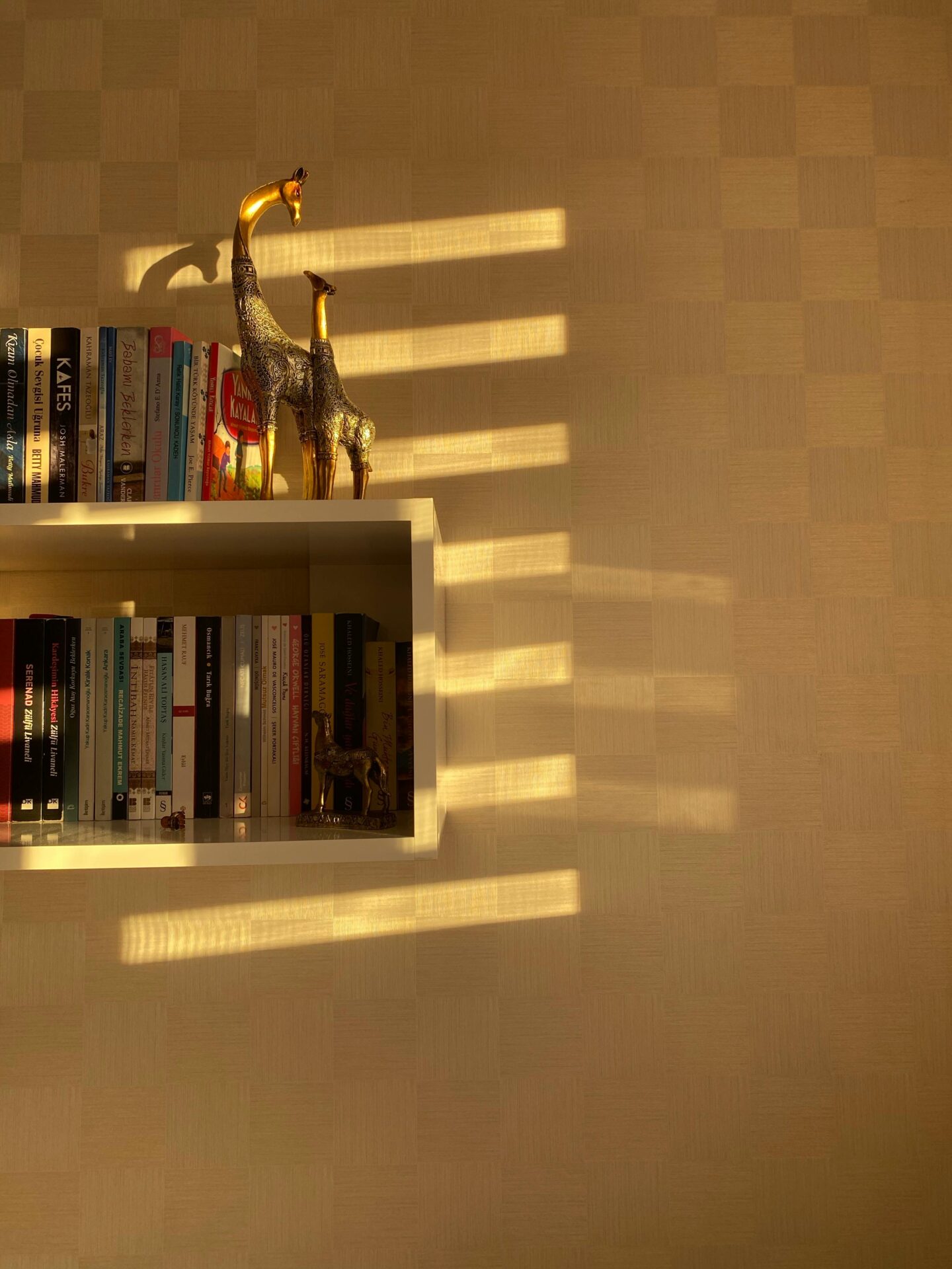 A white wall-mounted bookshelf filled with books and gold giraffe figurines, illuminated by warm sunlight casting horizontal shadows across a textured beige wall.