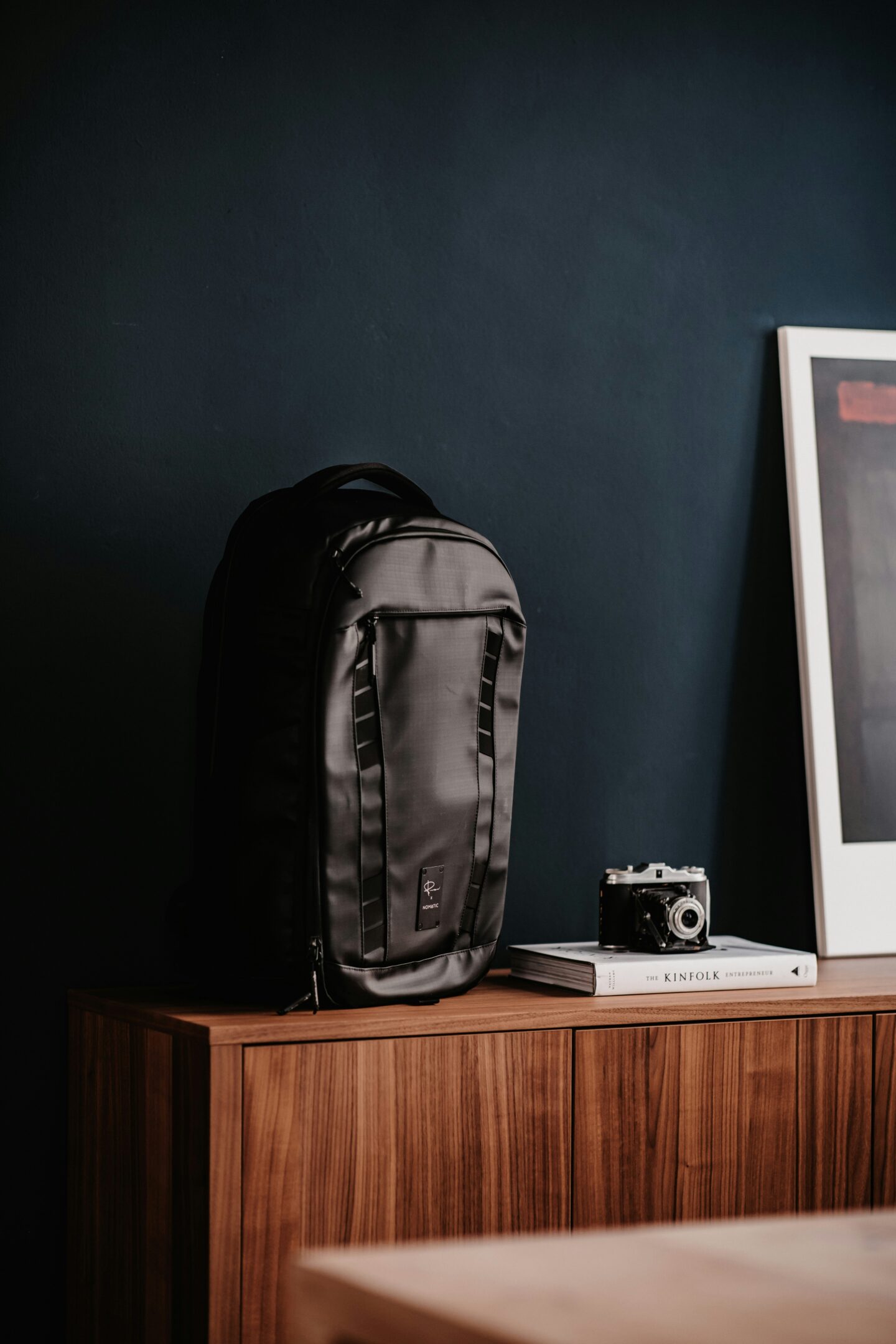 A minimalist interior scene featuring a black travel backpack on a wooden cabinet, next to a vintage camera placed on top of stacked books, with a dark blue wall and framed artwork in the background.