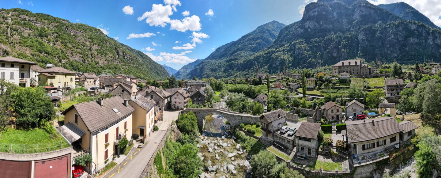 Drone view of the village of Giornico in Switzerland, showing stone houses, a river with a small bridge, and surrounding green mountains.
