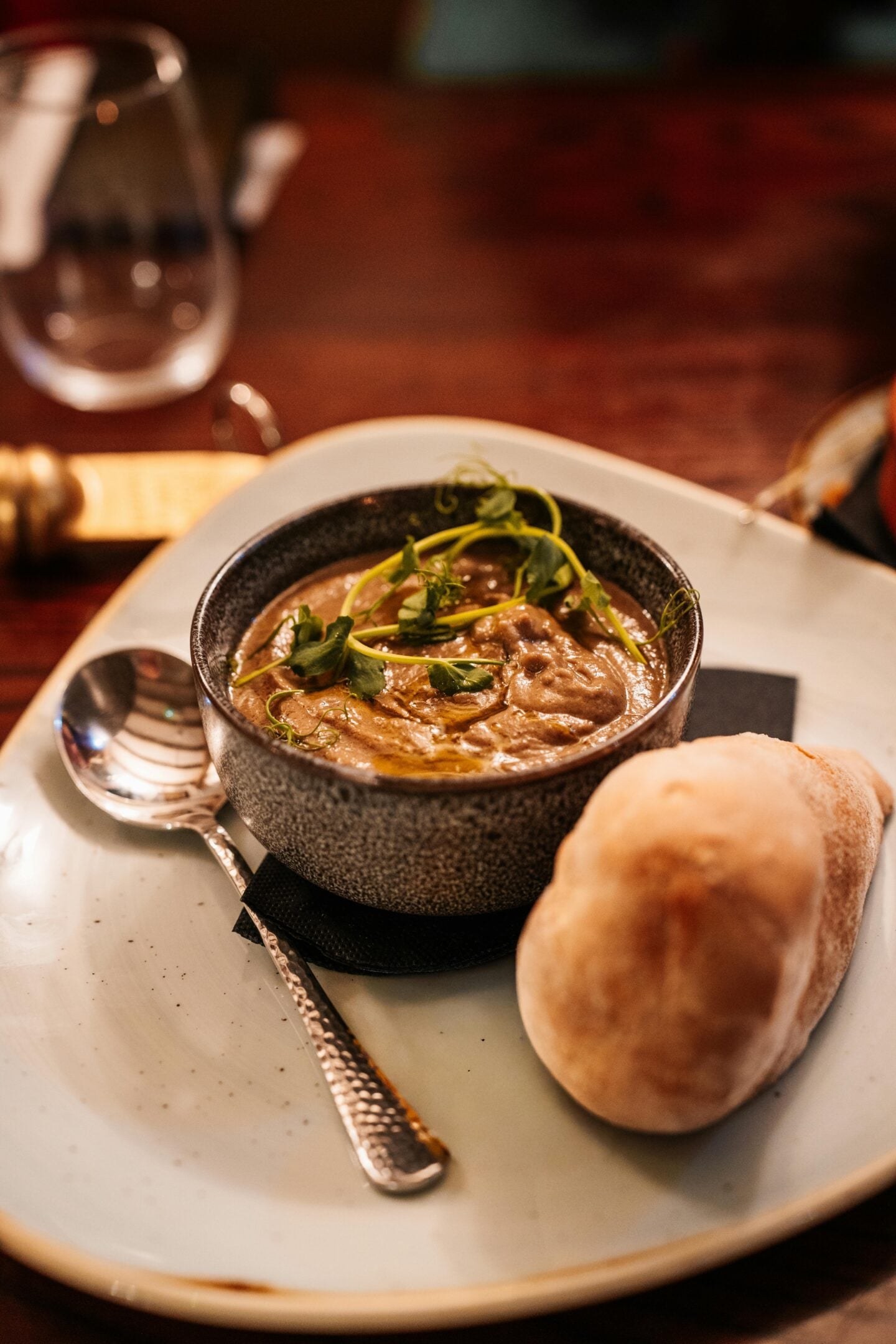 Bowl of creamy soup garnished with herbs, served with a bread roll on a white plate in a cozy restaurant setting.
