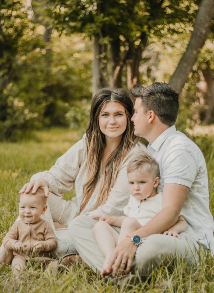 A family of four sitting on grass in a sunlit park, with two adults seated closely together and two young children sitting in front of them, surrounded by green trees and a soft natural background.