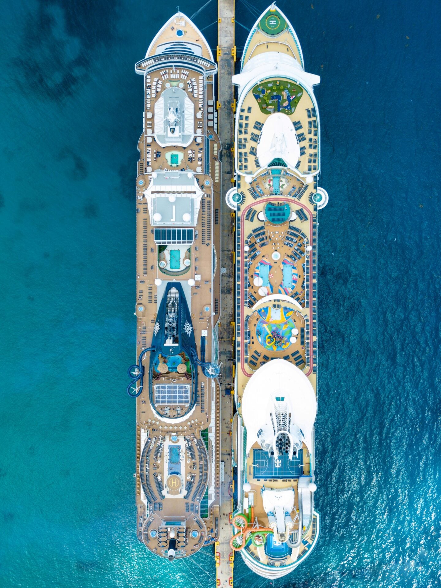 Aerial, top-down view of two large cruise ships docked side by side at a pier, showing detailed decks with pools, water slides, sun loungers, and recreational areas, surrounded by clear blue ocean water.