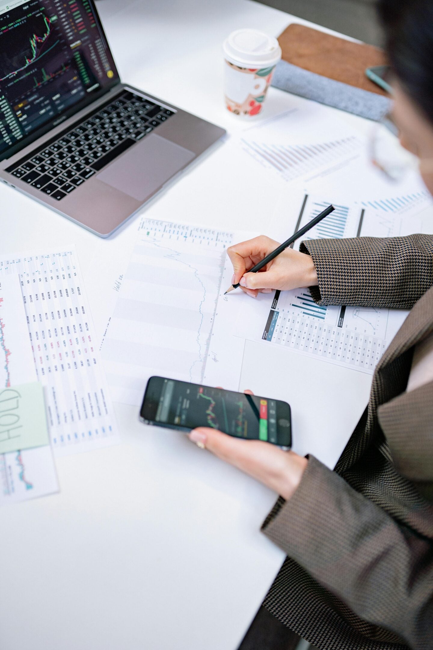 Person analyzing financial charts and stock market data on a laptop and smartphone while reviewing printed graphs and reports at a desk, illustrating modern financial planning and investment analysis.