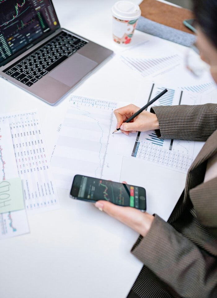Person analyzing financial charts and stock market data on a laptop and smartphone while reviewing printed graphs and reports at a desk, illustrating modern financial planning and investment analysis.